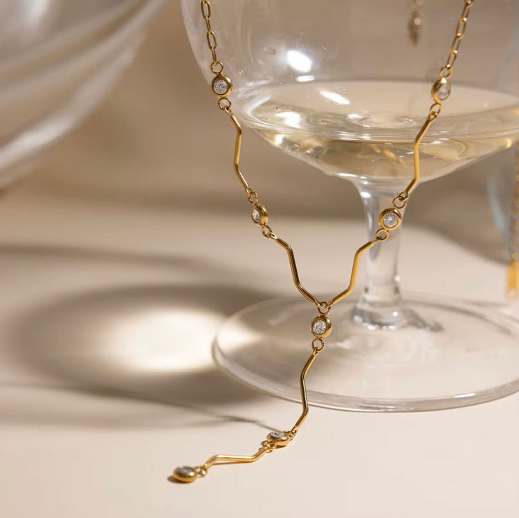Saya gold Y necklace with crystal accents draped over a glass coupe on a neutral background
