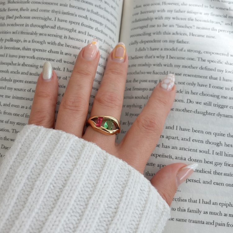 Close-up of Alissa gold ring with green and pink stones held in hand over open book