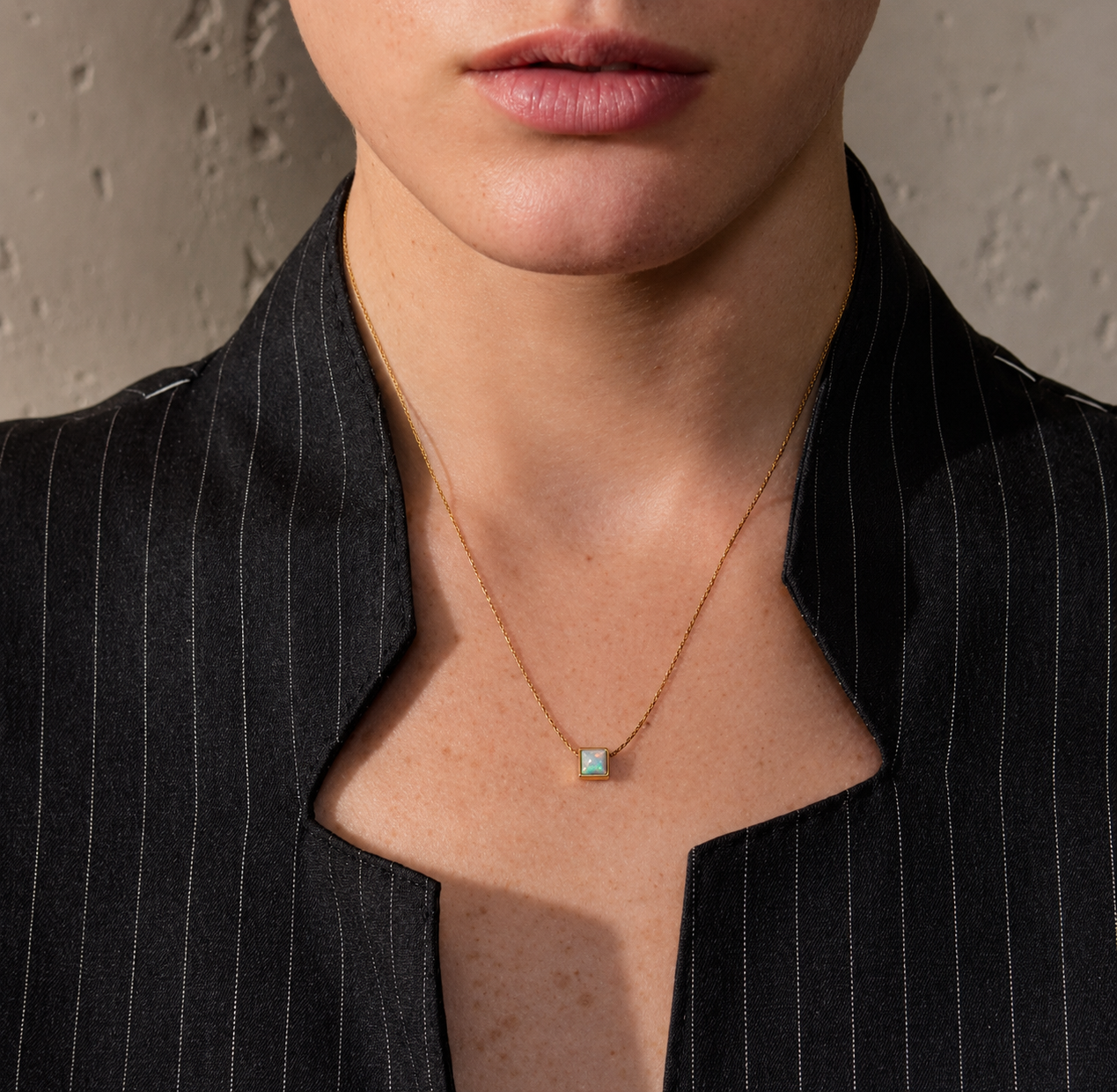 A close-up of a model wearing a delicate gold chain necklace with a small square pendant featuring a light blue-green stone, styled with a black pinstripe blazer.