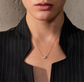 A close-up of a model wearing a delicate gold chain necklace with a small square pendant featuring a light blue-green stone, styled with a black pinstripe blazer.