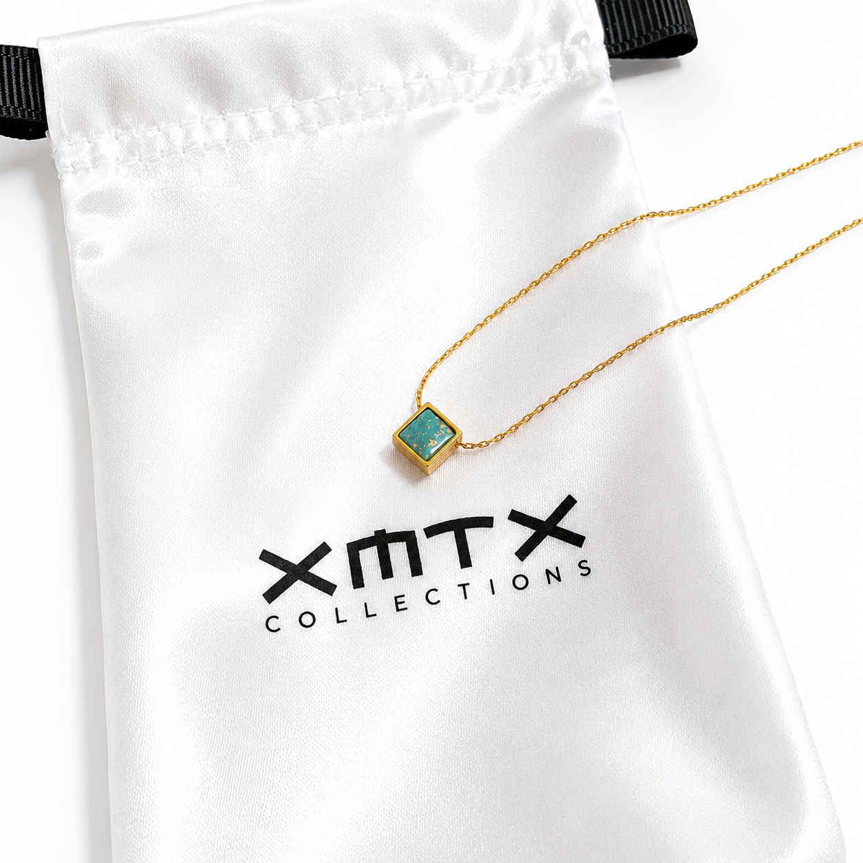A gold chain necklace with a square pendant featuring a green stone, displayed on a white satin jewelry pouch with the brand name &