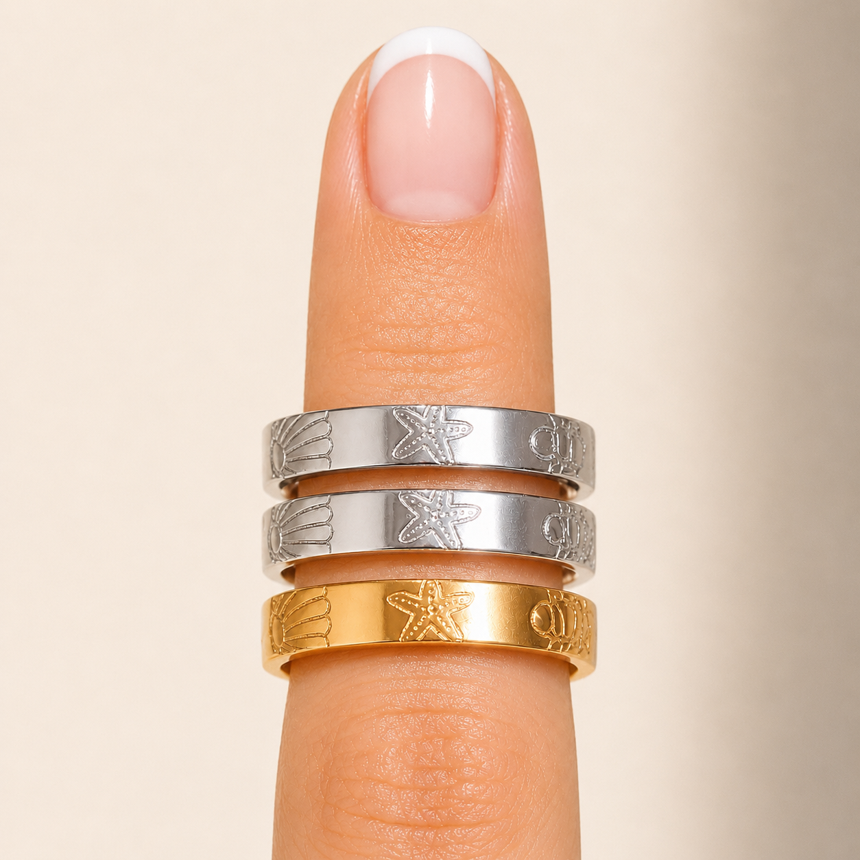 Three engraved rings stacked on a finger: two silver rings with starfish motifs and one gold ring, shown against a soft beige background.