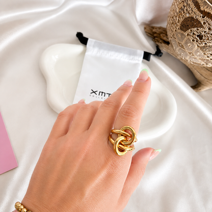 A close-up of a hand wearing a chunky gold ring with an interlocking knot design, resting on a white satin fabric background with a white drawstring pouch and decorative accessories.