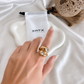 A hand wearing a gold ring with two clear teardrop-shaped gemstones, resting over a white XMTX collection drawstring pouch on a white satin fabric background.
