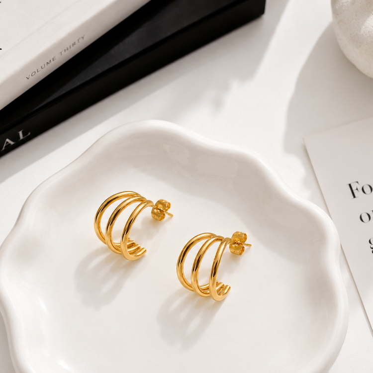 Shanti waterproof triple gold hoop earrings styled in a minimal luxury flat lay, featuring three sculptural gold bands.
