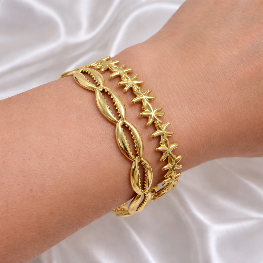 Gold cowrie shell bracelet and gold starfish bracelet stacked on a wrist against a white satin background