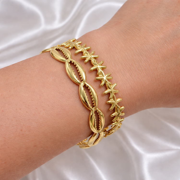 Gold cowrie shell bracelet and gold starfish bracelet stacked on a wrist against a white satin background